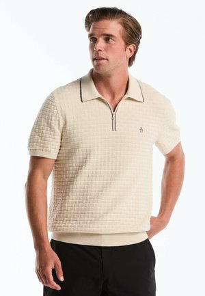Polo à manches courtes en crème, avec un motif texturé, un col zippé à mi-hauteur avec une bordure noire, et un emblème de pingouin sur la poitrine.