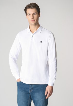 Polo a maniche lunghe bianco realizzato in cotone, con un colletto a tre bottoni e un piccolo logo nero sul lato sinistro del petto. Indossato con jeans blu.