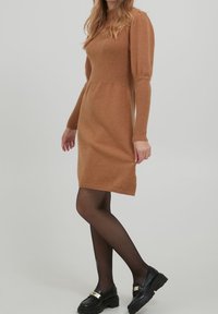 Abito a maglia in un caldo marrone con un corpetto aderente e maniche a sbuffo. Tessuto testurizzato, lunghezza a metà coscia, abbinato a scarpe nere e collant.