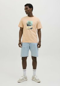 Camiseta de algodón color durazno con diseño gráfico de palmera, combinada con shorts de mezclilla azul claro. El modelo lleva zapatillas blancas y calcetines con logo.