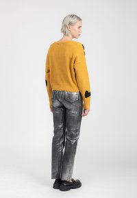 Vrouw met kort blond haar die een mosterdkleurige trui met zwarte hartpatches en metallic zilveren broeken draagt, staande met haar gezicht naar rechts tegen een witte achtergrond.