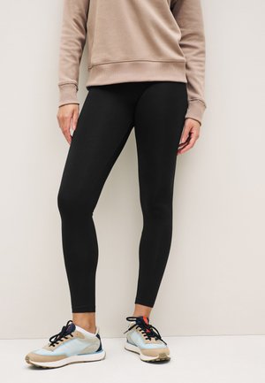 Person iført sorte leggings, beige sweatshirt og hvide sneakers med marineblå og orange accenter, stående mod en ensfarvet lys væg.