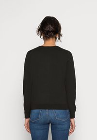 Pull noir à manches longues avec un col rond et une texture côtelée, porté avec un jean bleu. La coupe est décontractée avec un dos droit.