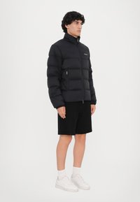 Sort puffersjakke med højt krave, lynlåslukning foran og sidelommer. Har quiltede sektioner til isolering og et minimalistisk design.