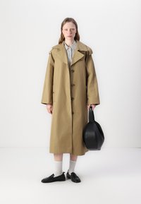 Beige trenchcoat laget av lett stoff, med dobbelspent front, bred krage og store sidelommer. Svart håndveske og sko.