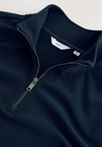 Námořnicky modrý fleecový pulovr s vysokým límcem, zapínáním na ¼ zip a hladkou texturou. Obsahuje kovové zipy a viditelnou značkovou visačku.