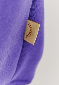 Fialový oděv s jemnou texturou; na boku je malá, hnědá etiketa s designem slunečního paprsku.