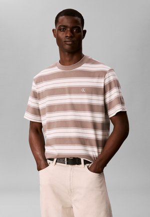 Homme portant une chemise à manches courtes beige et blanche rayée avec le logo "cK", mains dans les poches, pantalon beige et ceinture noire, debout devant un fond gris.