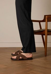 Infradito marroni con cinturini incrociati e suola piatta, indossati con pantaloni neri, su un pavimento di legno accanto a una sedia.