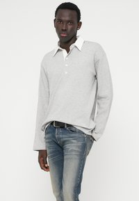 Pólo cinza de mangas longas com colarinho branco e três botões, combinado com jeans azuis desbotados. O tecido parece suave e texturizado.