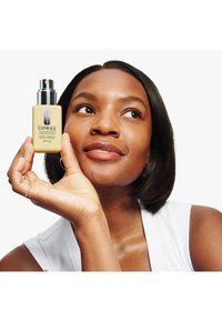 Clinique fuktighetskrem i en gul flaske med en sølvpumpe, merket SPF 50, holdt av en hånd mot en hvit bakgrunn.