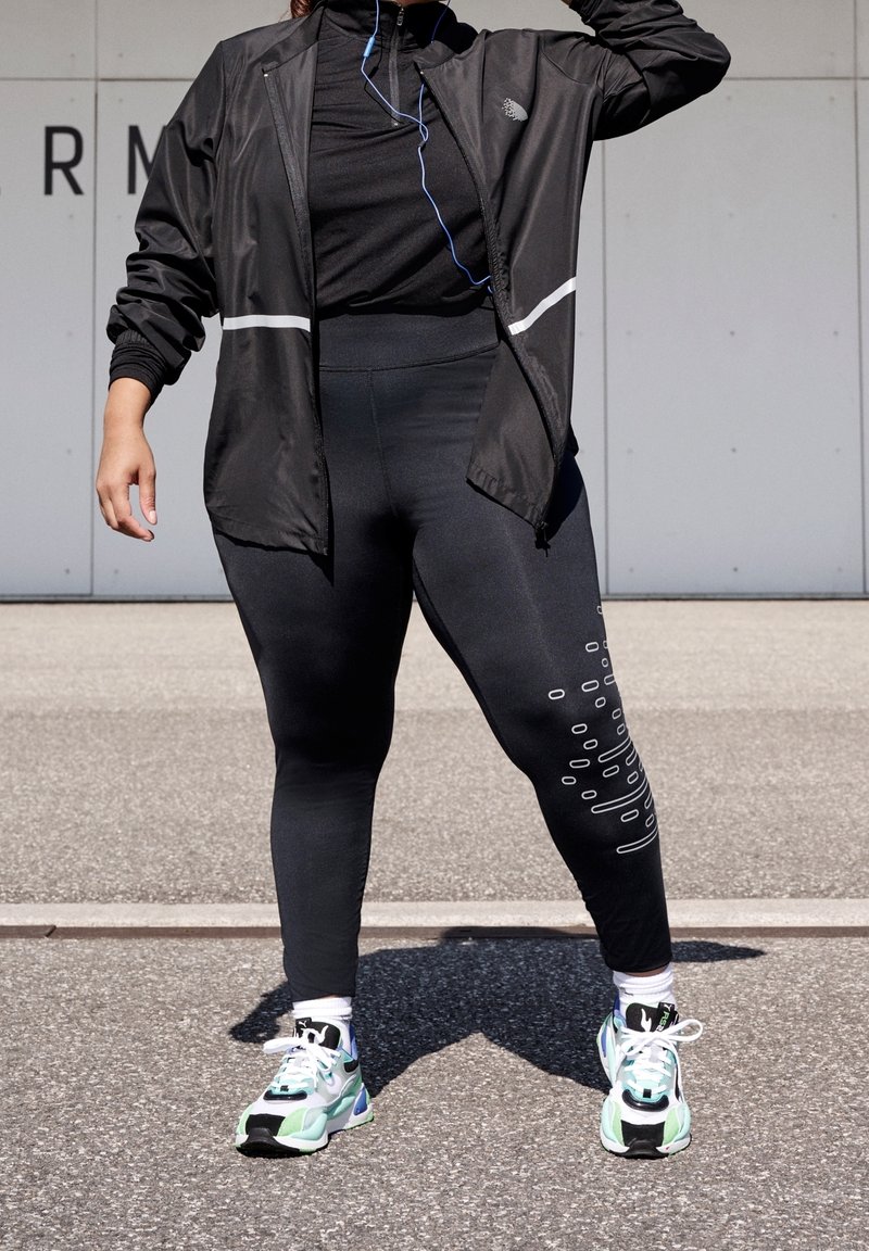 Personne portant une veste de sport noire et des leggings avec des baskets blanches et vertes, debout sur le trottoir à l'extérieur.