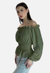 Olivgrüne schulterfreie Bluse mit Rüsche am Hals, Puffärmeln und gerafftem Taillendetails, kombiniert mit hellblauen Jeans.