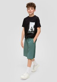 Schwarzes Grafik-T-Shirt mit einem fotografischen Druck und Text. Kombiniert mit lockeren grünen Shorts und weißen Turnschuhen mit sichtbaren Schnürsenkeln.