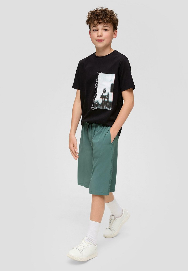 Schwarzes Grafik-T-Shirt mit einem fotografischen Druck und Text. Kombiniert mit lockeren grünen Shorts und weißen Turnschuhen mit sichtbaren Schnürsenkeln.
