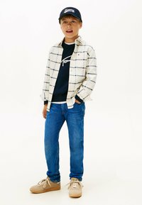 Dreng iført en hvid og marineblå ternet skjorte over en marineblå sweatshirt, blå jeans og beige sneakers med creme farvede snørebånd, afsluttet med en marineblå kasket.