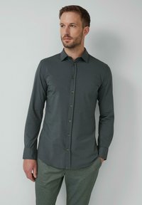 Mann mit kurzem Haar und Bart, der ein dunkelgraues, knöpfbares Hemd und eine hellgraue Hose trägt, steht vor einer schlichten, hellen Wand.