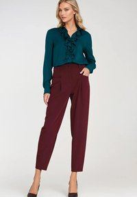 Zielona bluzka w kolorze szmaragdowym z falbaną z przodu, zestawiona z wysokim stanem bordowymi spodniami. Ubranie charakteryzuje się gładką teksturą i dopasowanym krojem.