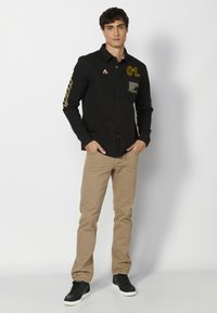 Camisa negra de botones con gráficos amarillos, un bolsillo en el pecho y mangas largas. Combinada con pantalones beige de corte ajustado y zapatillas negras.