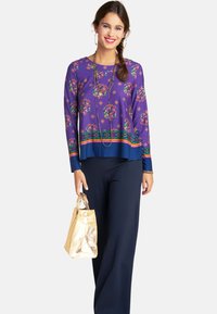 Lila Langarmoberteil mit Blumenmuster und gestreiftem Saum, kombiniert mit marineblauen weit geschnittenen Hosen und einer metallisch goldenen Handtasche.