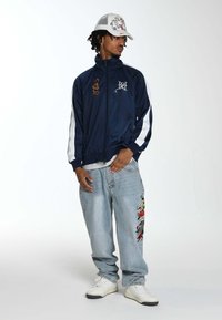 Marineblauwe zip-up jack met witte zijstrepen en borduurwerk. Lichtblauwe jeans met kleurrijk zijborduurwerk. Witte sneakers en een bedrukte pet.