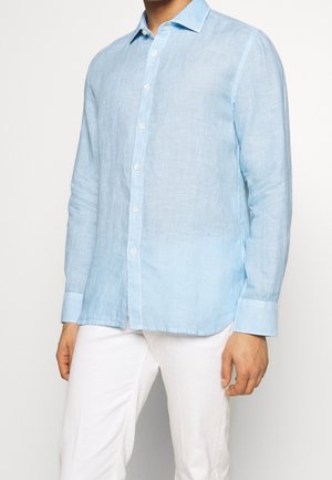 Mężczyzna ubrany w jasnoniebieską lnianą koszulę z długim rękawem, białymi guzikami i białe spodnie, stojący na jednolitym tle.