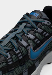 Αθλητικό παπούτσι Nike με σκούρο πράσινο πλεκτό πάνω μέρος, γκρι και μαύρες λεπτομέρειες. Μπλε σήμα Swoosh και θηλιές κορδονιών με υφή στην επιφάνεια.