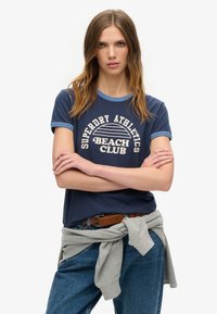 Granatowa koszulka z jasnoniebieskimi wykończeniami, z białym napisem graficznym "Superdry Athletics Beach Club." Połączona z wysokim stanem dżinsowymi spodniami i szarym swetrem.