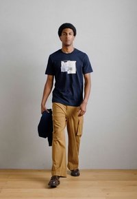 T-shirt azul-marinho com estampa gráfica, combinada com calças cargo bege. O modelo segura um blazer azul, está de pé sobre um chão de madeira contra uma parede neutra.