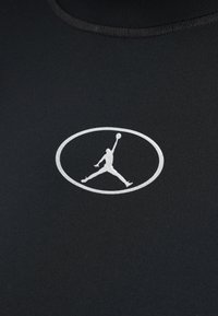 Juodas sportinis marškinėlis su ryškiu baltu Jordan logotipu ovalo formoje. Medžiaga yra lygi ir šiek tiek elastinga.