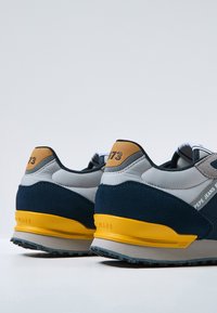 Baskets bleu marine et gris avec semelle jaune. Tige en daim et maille, col rembourré, système de laçage, logo au niveau du talon, design texturé sur le côté.