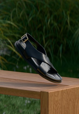 Melna ādas sandalete ar krusteniskām siksnām un slingback ar zelta sprādzi. Gluda tekstūra, plakans zolītes, izvietota uz koka virsmas.