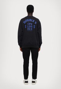 Svart genser med fet blå tekst på baksiden. Har rund hals, lange ermer og ribbede mansjetter. Kombinert med svarte bukser og sneakers.