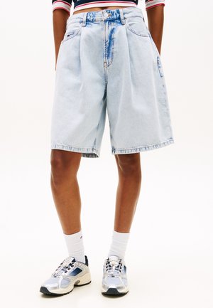 Persoană purtând pantaloni scurți largi din denim albastru deschis, șosete albe până la gleznă și încălțăminte sport argintie, stând în picioare pe un fundal alb simplu.