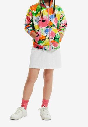 Felpa con zip colorata con un design floreale in rosa, giallo e verde. Indossata sopra una gonna bianca con sneakers bianche e calzini rosa.