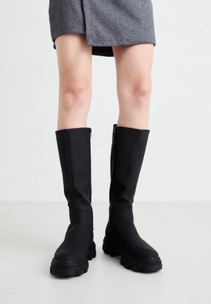 Jambes portant une jupe grise et des bottes noires montantes avec des semelles épaisses, debout sur un sol clair devant un fond uni.