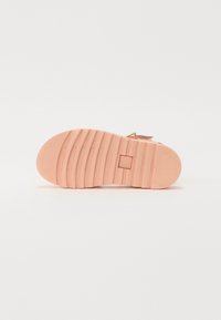 Sandala z teksturirano, roza gumijasto podplatom z vodoravnimi utori. Prilagodljiv trak z kovinskim dodatkom in mehkim zgornjim delom iz blaga.