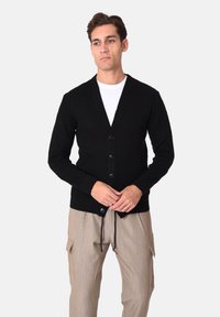 Cardigan nero a coste con collo a scialle, caratterizzato da cinque bottoni sul davanti e maniche lunghe. Indossato con pantaloni cargo di colore chiaro.
