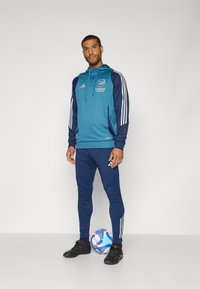 Teal og marineblå atletisk træningsdragt med tre hvide striber på ærmerne og siden, med et Arsenal-logo, parret med sorte sportssko og en fodbold.