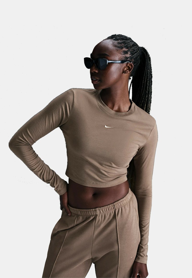 Crop top marrone a maniche lunghe con scollo rotondo, caratterizzato da un piccolo logo Nike bianco, abbinato a pantaloni sportivi marroni coordinati.