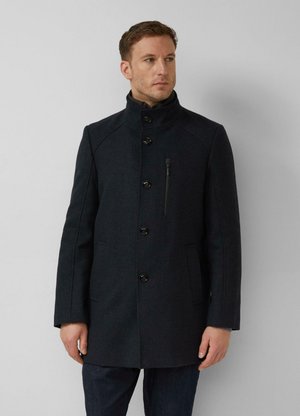 Man draagt een donker wollen jas met knopen, een hoge kraag en een borstzak met rits, staand tegen een effen lichte achtergrond.
