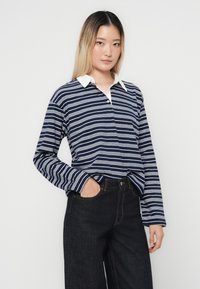 Marineblå langermet polo skjorte med hvite horisontale striper, hvit krage og en avslappet passform, kombinert med mørk denimbukser.