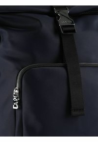 Približen posnetek temnopoltega žepka na torbi s srebrnim zadrgo in črnim trakom s srebrno zaponko z napisom "BOGNER".