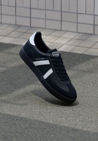 Schwarz-weißer Jack & Jones Sneaker mit Schnürsenkeln, weißen Streifen und strukturierten Sohle auf grauem Pflaster neben einem gefliesten Wandhintergrund.