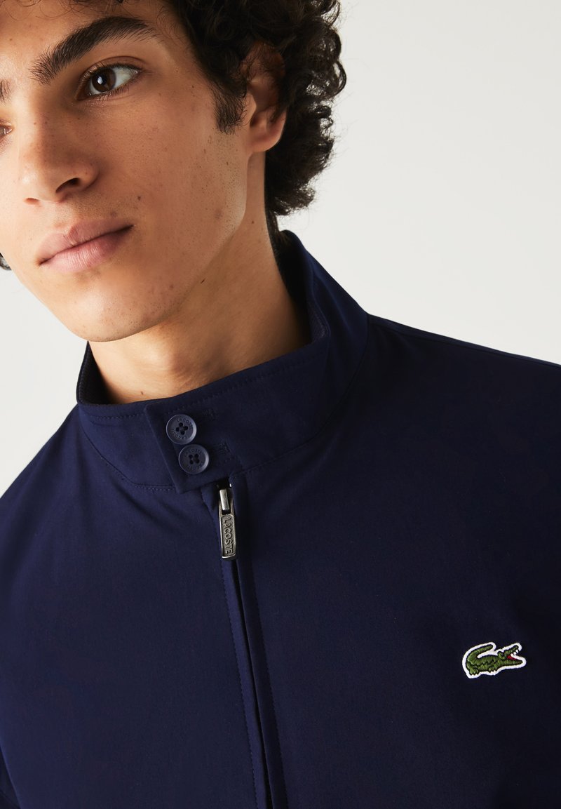 Jeune homme portant une veste zippée bleu marine avec un col montant et un logo de crocodile vert sur la poitrine.