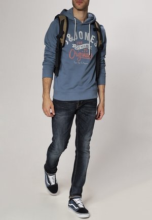 Uomo che cammina indossando una felpa blu con cappuccio "J&Jones", jeans scuri, sneakers nere e bianche, e che porta uno zaino marrone su uno sfondo chiaro.