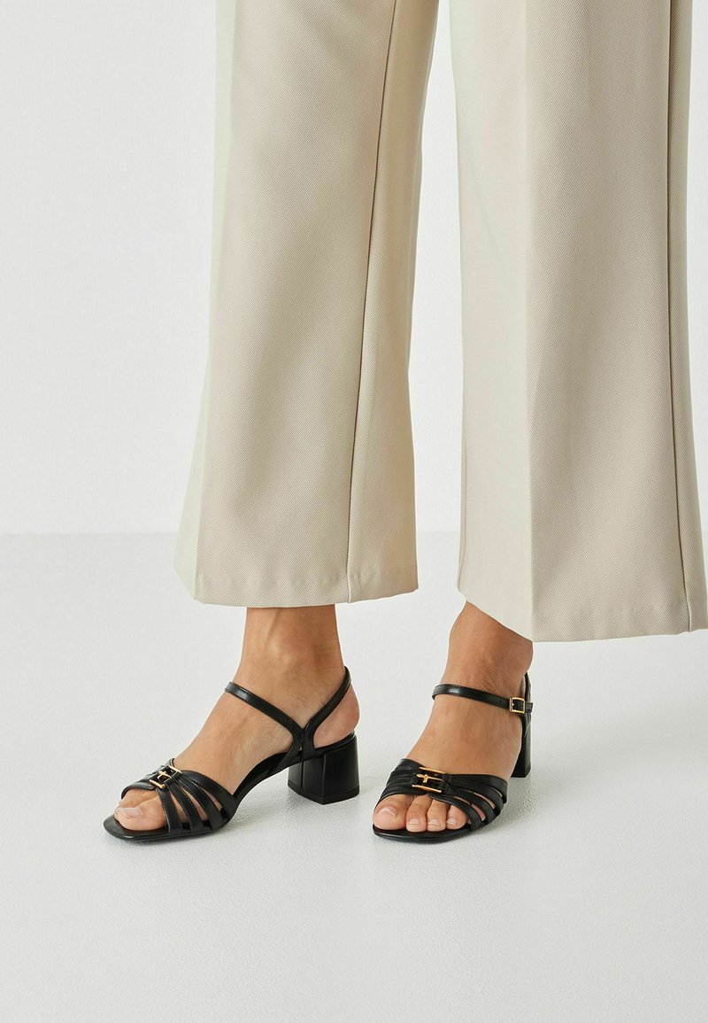 Črne sandale z naramnicami in blok peto imajo minimalističen dizajn, zlat poudarek na okovju in odprt sprednji del. Oblečene so z bež širokimi hlačami.