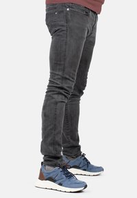 Jeans oscuros de corte slim con un ligero desgaste, combinados con zapatillas azules que tienen acentos en negro y marrón y una suela texturizada.