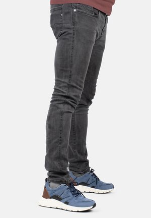 Jeans slim-fit grigio scuro con leggera scoloritura, abbinati a sneakers blu con accenti neri e marroni e suola testurizzata.