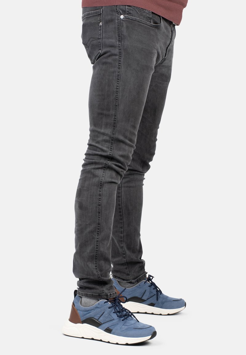 Jeans oscuros de corte slim con un ligero desgaste, combinados con zapatillas azules que tienen acentos en negro y marrón y una suela texturizada.